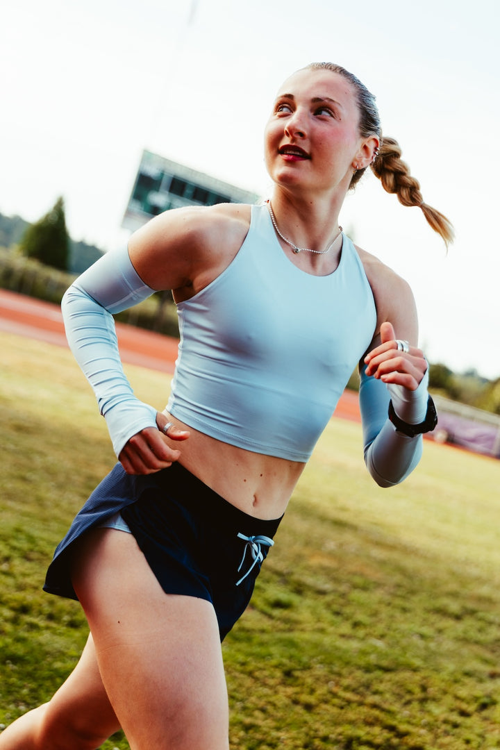 A woman running on an outdoor track wearing a light blue athletic crop top, dark navy shorts, and a light blue arm sleeve, captured in motion against a blurred grassy field background.